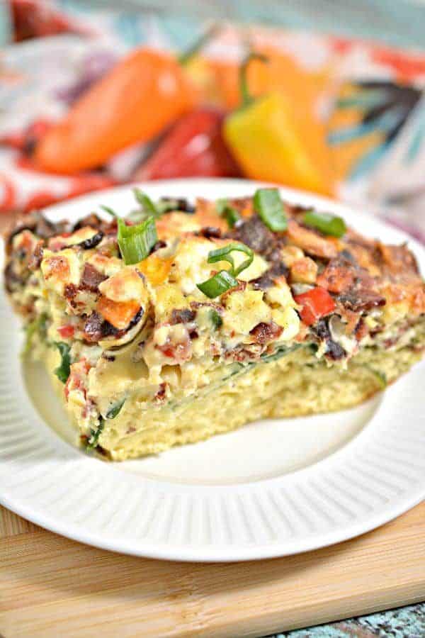 Close up of slice of breakfast casserole with chopped green onions on top as garnish, mini bell peppers in background