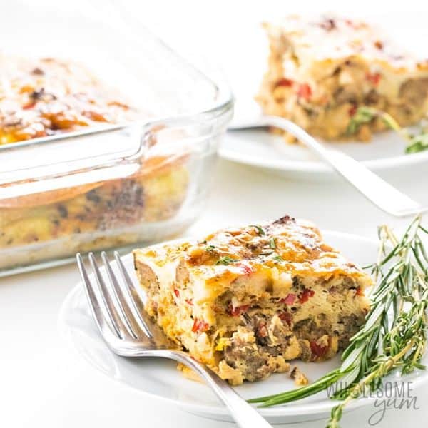 Slice of breakfast casserole on plate with fork and rosemary, casserole dish in background next to another plated piece of casserole