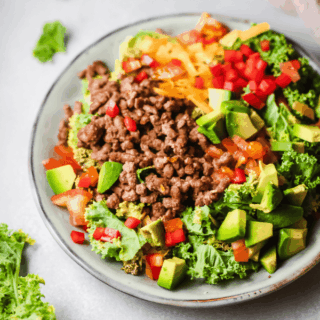 A plate of keto taco salad made with ground beef, avocado, chopped bell peppers, shredded cheddar cheese, and mixed greens.
