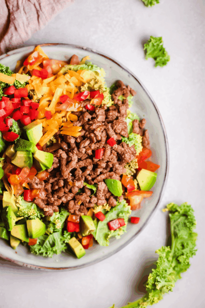 Bright and colorful keto taco salad bowl filled with ground beef, chopped avocado, shredded cheese, and mixed greens on a light surface.
