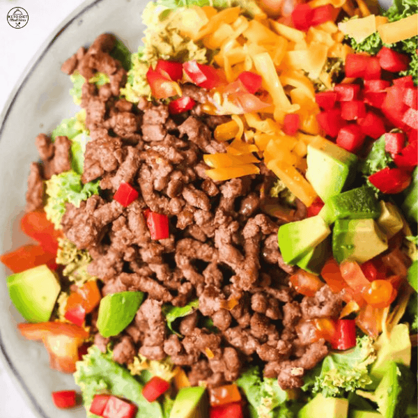 A fresh taco salad with seasoned ground beef, avocado, shredded cheese, diced red bell peppers, and lettuce served in a bowl.