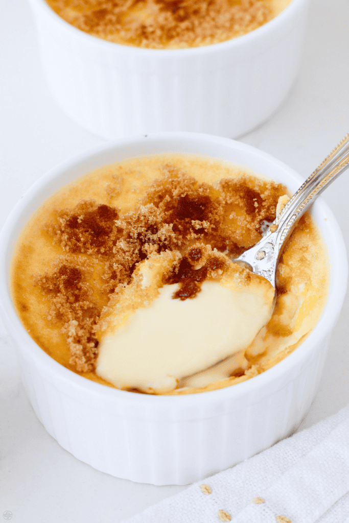A white ramekin filled with smooth keto creme brulee topped with caramelized sweetener, with a spoon lifting a creamy bite.