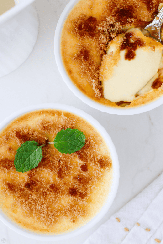 Overhead view of two keto creme brulees in white ramekins, one garnished with mint and the other with a spoonful scooped out.