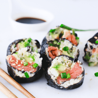 Close-up of several keto sushi rolls arranged on a white board, garnished with green onion and sesame seeds next to soy sauce.