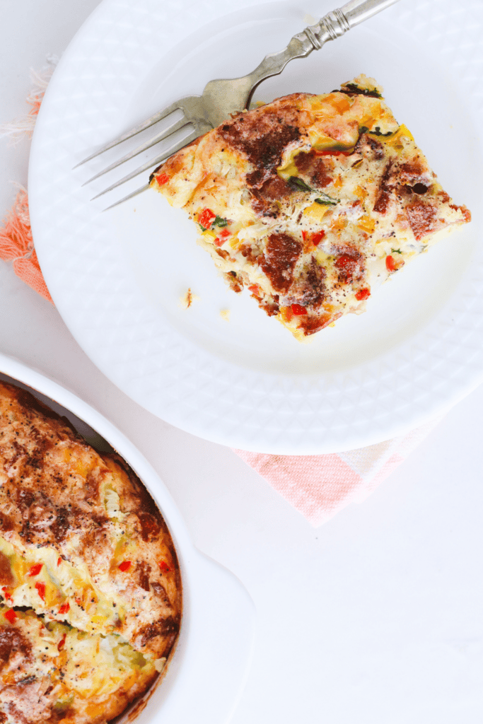 A plated slice of spinach and feta egg bake with colorful vegetables and a silver fork on a white plate.