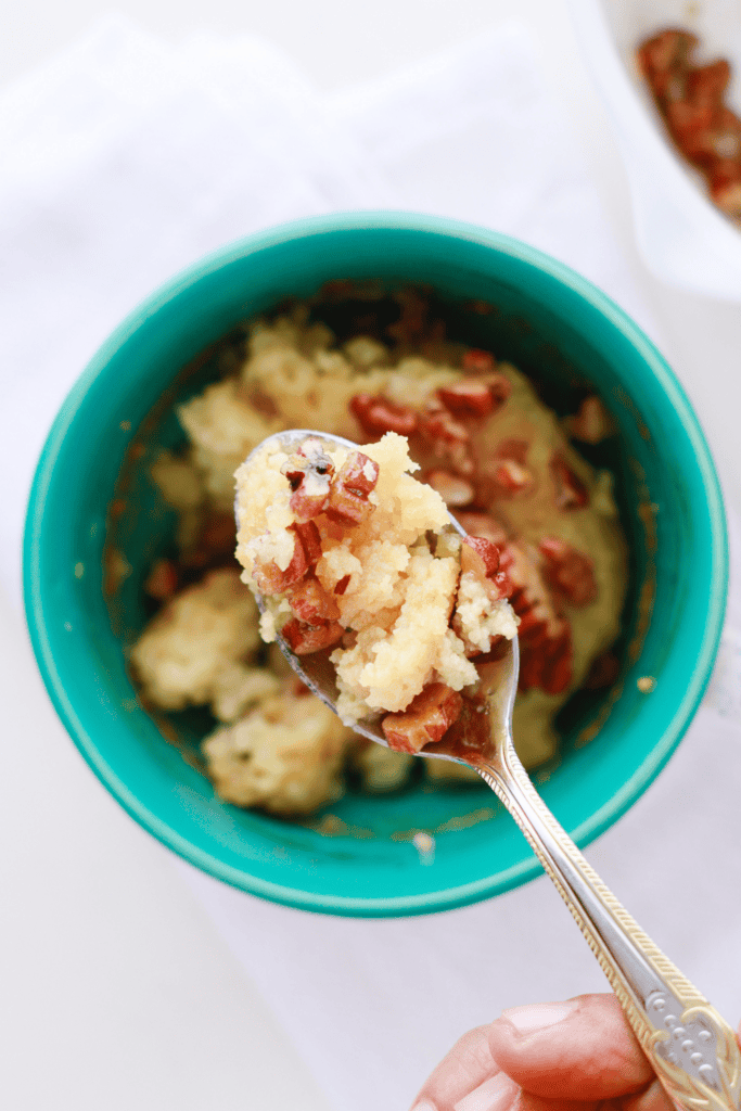 A spoonful of fluffy keto pecan pie mug cake topped with buttery chopped pecans, held over a teal mug.