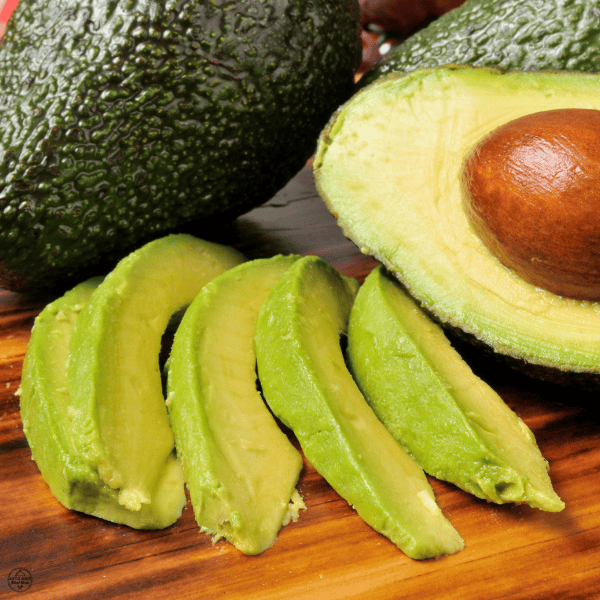Fresh avocado slices and a halved avocado with the pit displayed on a wooden board.