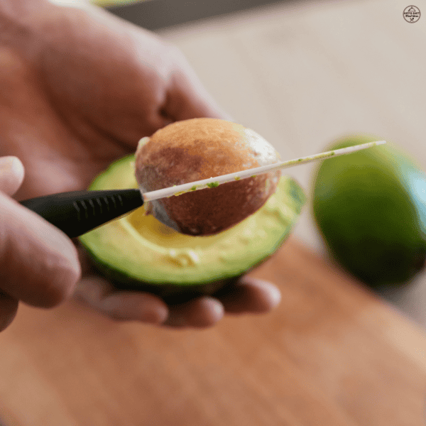 A person removing the pit from a halved avocado using a knife over a kitchen counter.